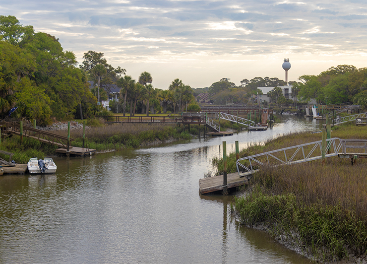 South Carolina Islands - A photograph of various docks all leading into a stretch of water, possibly a river. Green trees dot the sides of the river. The sky is overcast with some clouds.