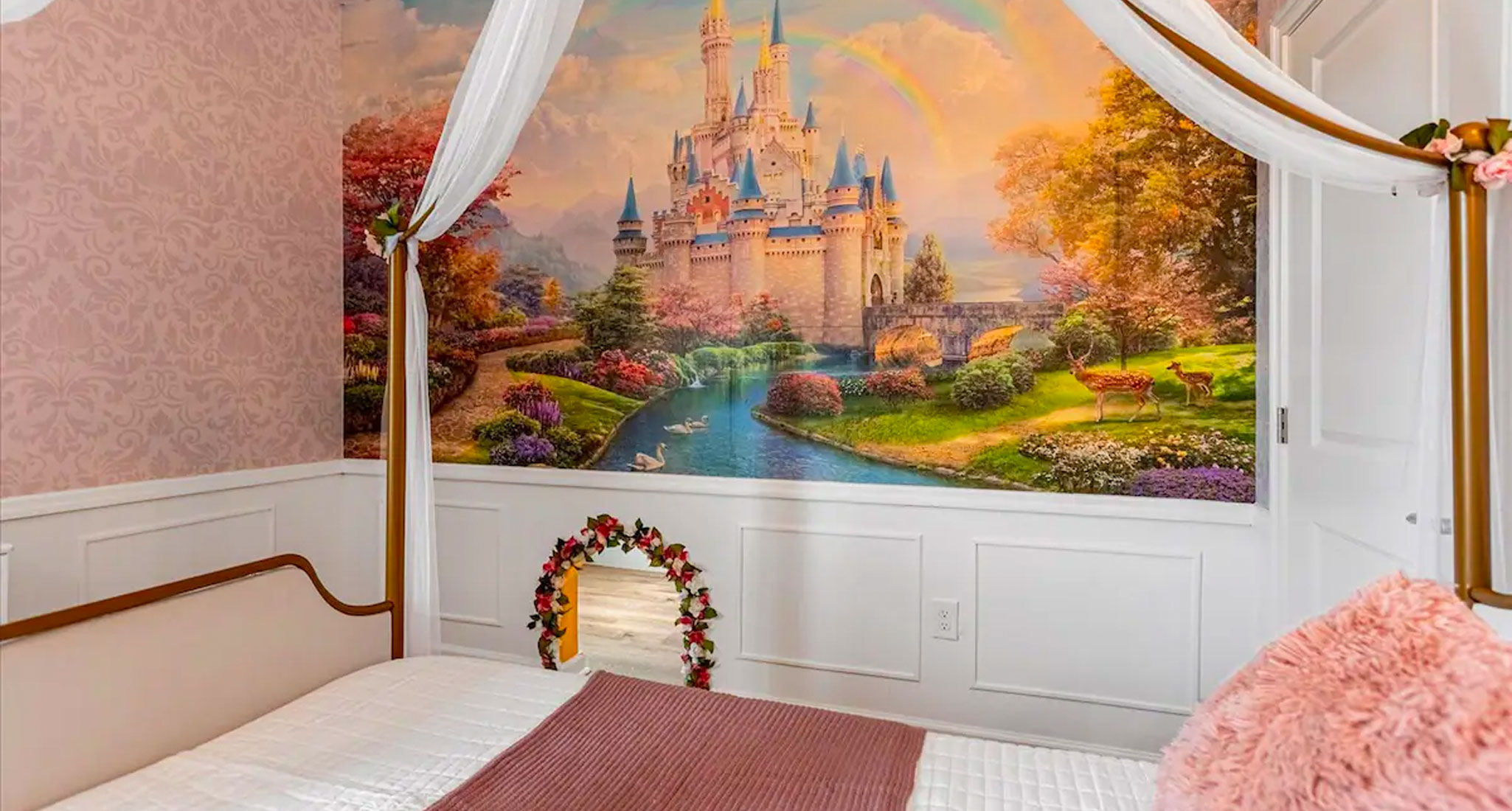 best-kid-friendly-airbnbs-in-orlando: A photograph of a canopy bed with a white duvet and pink pillows. Behind it is a mural of Cinderella's castle. There is also a small doggie door framed with flowers. The walls of the room are pink.
